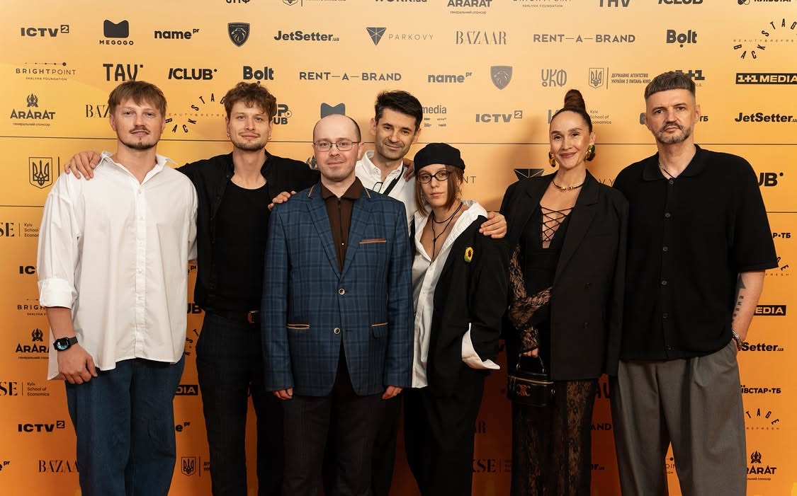 The film crew, actors, and director are standing in front of a banner at the film's premiere